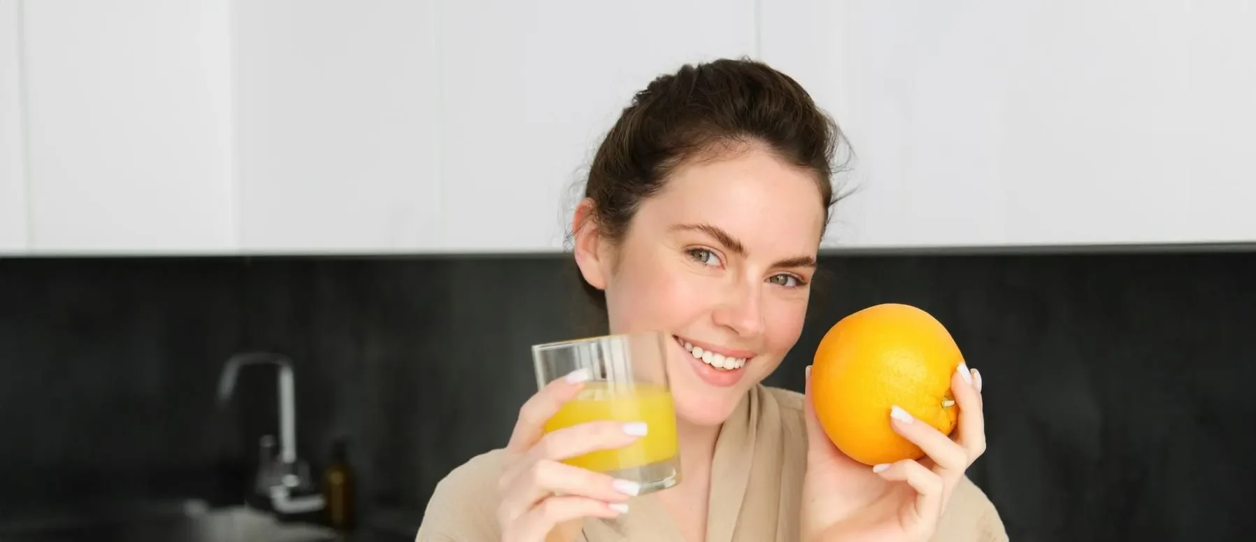 Image of goodlooking healthy woman in bathrobe drinking fresh juice showing orange fruit posing in